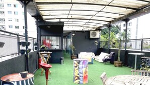 An outdoor patio area with green artificial turf, a round wooden table, and several chairs. - Hacker Hostel (São Paulo)