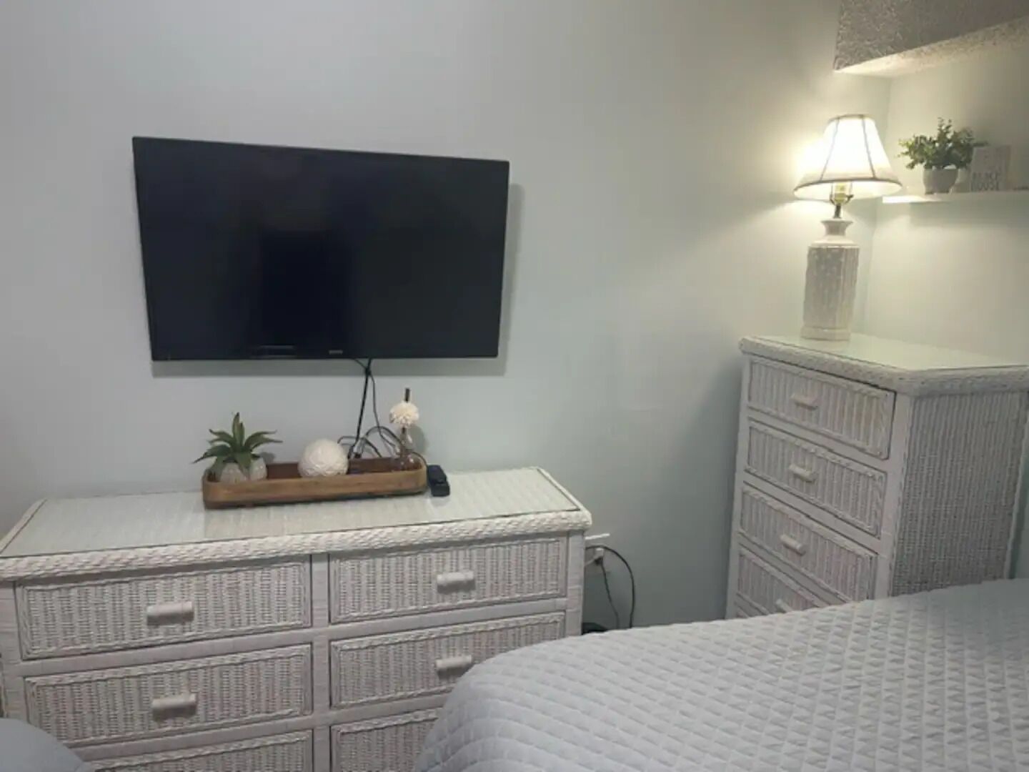 Un dormitorio con una televisión montada en la pared, una mesita de noche con lámpara y un aparador blanco con cajones.
