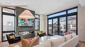 A modern living room with a large sofa, a glass coffee table, and a fireplace. There is a large window with a view of buildings outside. - One Riverfront Village by EWH (Avon)