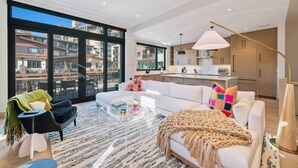 A modern apartment with a living room, kitchen, and large windows offering a city view. - One Riverfront Village by EWH (Avon)