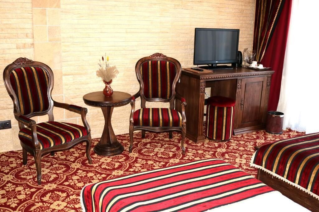 A hotel room with two upholstered chairs, a small round table with a vase, a television on a wooden desk, and a patterned carpet.