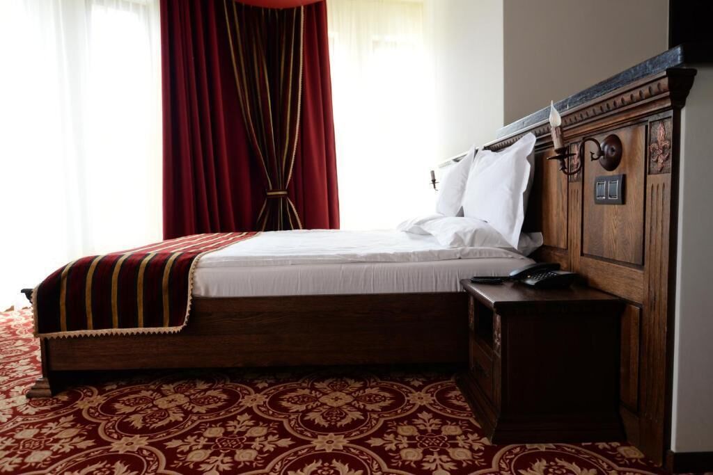 A hotel room with a wooden bed, a red patterned carpet, a wooden nightstand, and a red curtain.