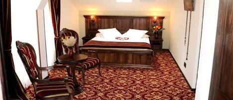 Un dormitorio con una cama grande, cabecera de madera, alfombra roja estampada, una mesita con un jarrón y una silla.