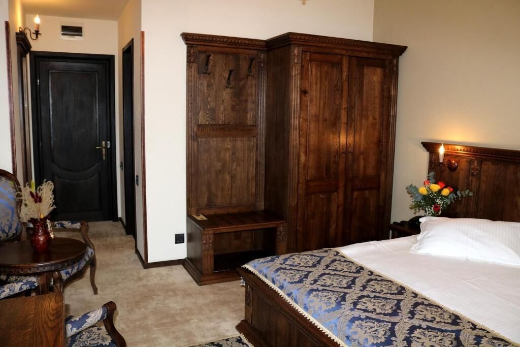 A bedroom with a large wooden wardrobe, a bed with patterned bedding, a small table with a vase, and a door with a light fixture.