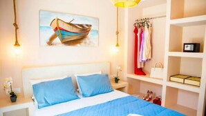 A hotel room with a bed, a painting of a boat, a wardrobe, and a shelf with items. - Vista Al Mar (Santorini)