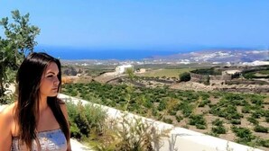 A view of a coastal landscape with a clear sky, a distant view of the sea, and a person standing on a balcony overlooking the scenery. - Vista Al Mar (Santorini)
