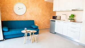 A modern living room with a blue sofa, a round table with a tray, a clock on the wall, and a kitchen area with cabinets and a microwave. - Vista Al Mar (Santorini)