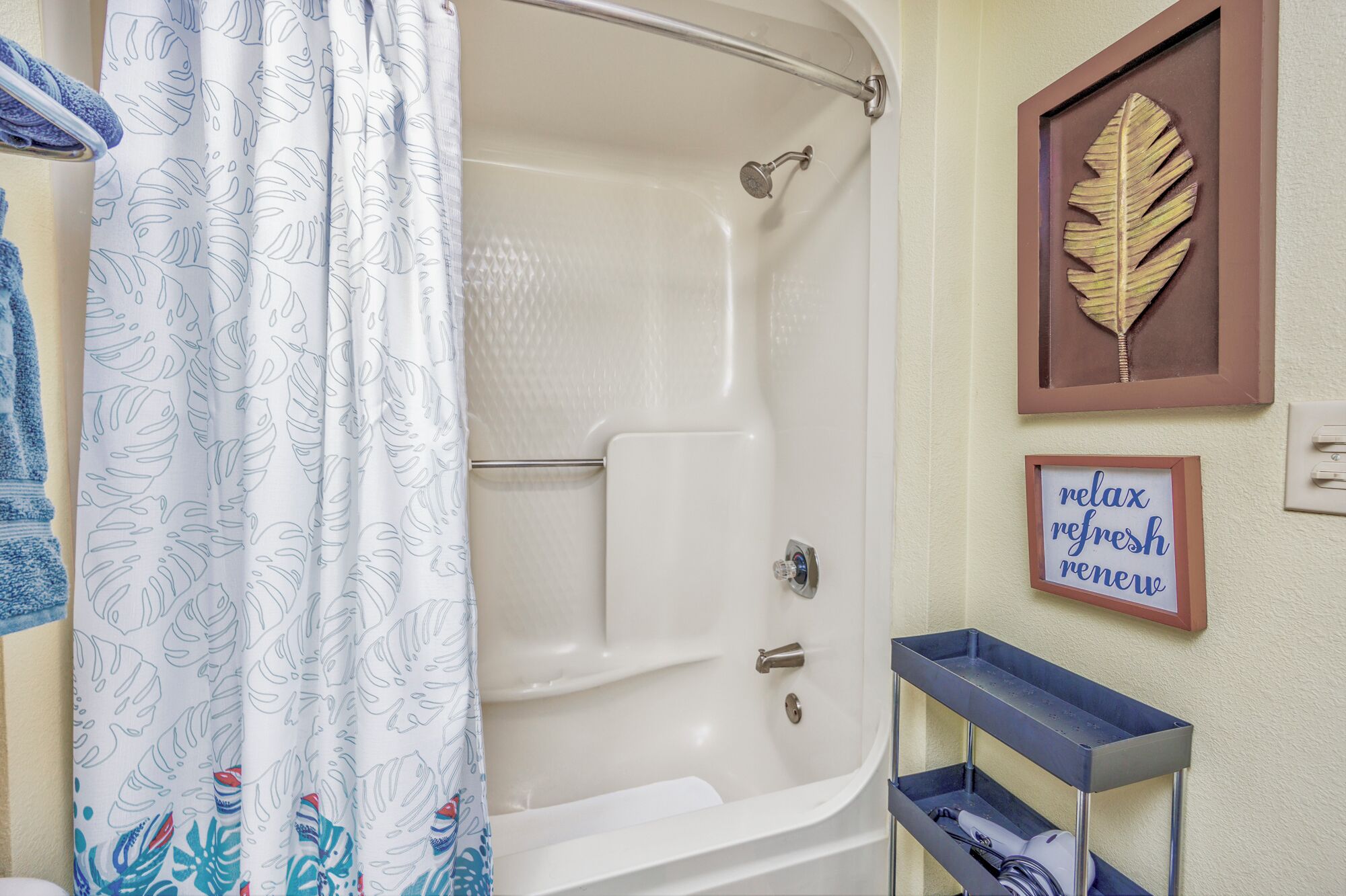 Ein Badezimmer mit Dusche, Handtuchhalter, einer Blattdekoration im Bilderrahmen und einem Regal mit dem Schriftzug "relax refresh renew".