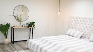 A bedroom with a tufted headboard, a round mirror, a bedside table with a clock and books, and two potted plants. - Modern cozy home, pet friendly (Wolcott)
