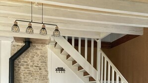 Uma área de refeições com uma mesa de madeira, um banco e uma cadeira. Há uma escada com corrimãos brancos e uma parede de pedra com uma placa.