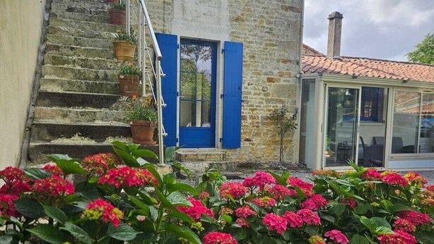 Uma casa de pedra com portadas azuis, um lance de escadas e um jardim com flores cor-de-rosa.