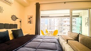 Une chambre moderne avec un grand lit, un canapé, une table à manger avec des chaises jaunes et une vue sur des immeubles de grande hauteur.