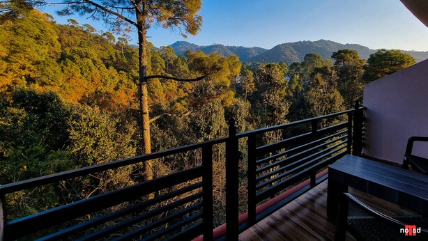 A balcony with a wooden table and chairs, overlooking a forested mountain range. - Nature Nest By Nomad Hotels (Dalhousie)