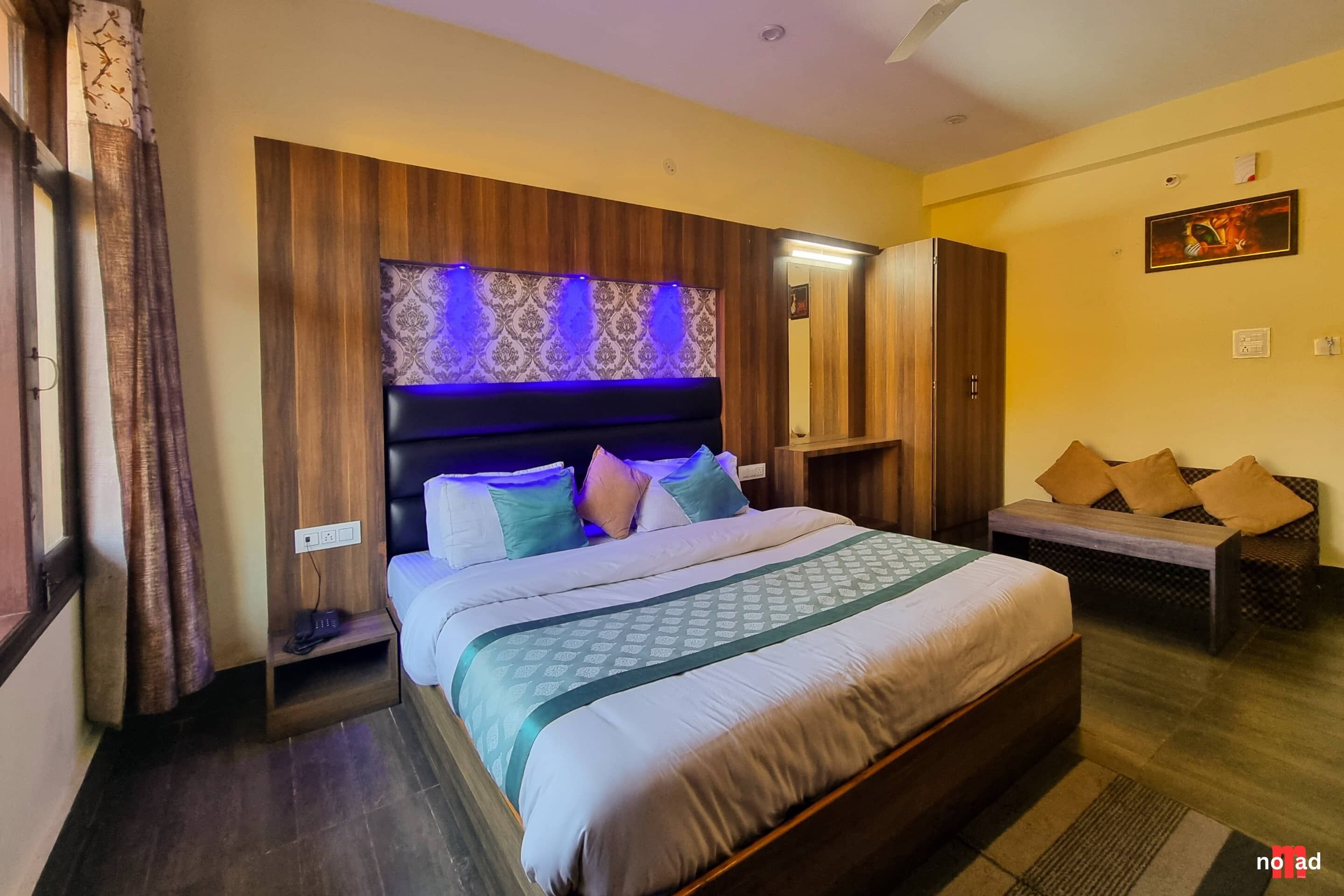 Habitación de hotel con una cama grande, cabecera de madera con iluminación azul, un banco con cojines y un espejo en la pared.