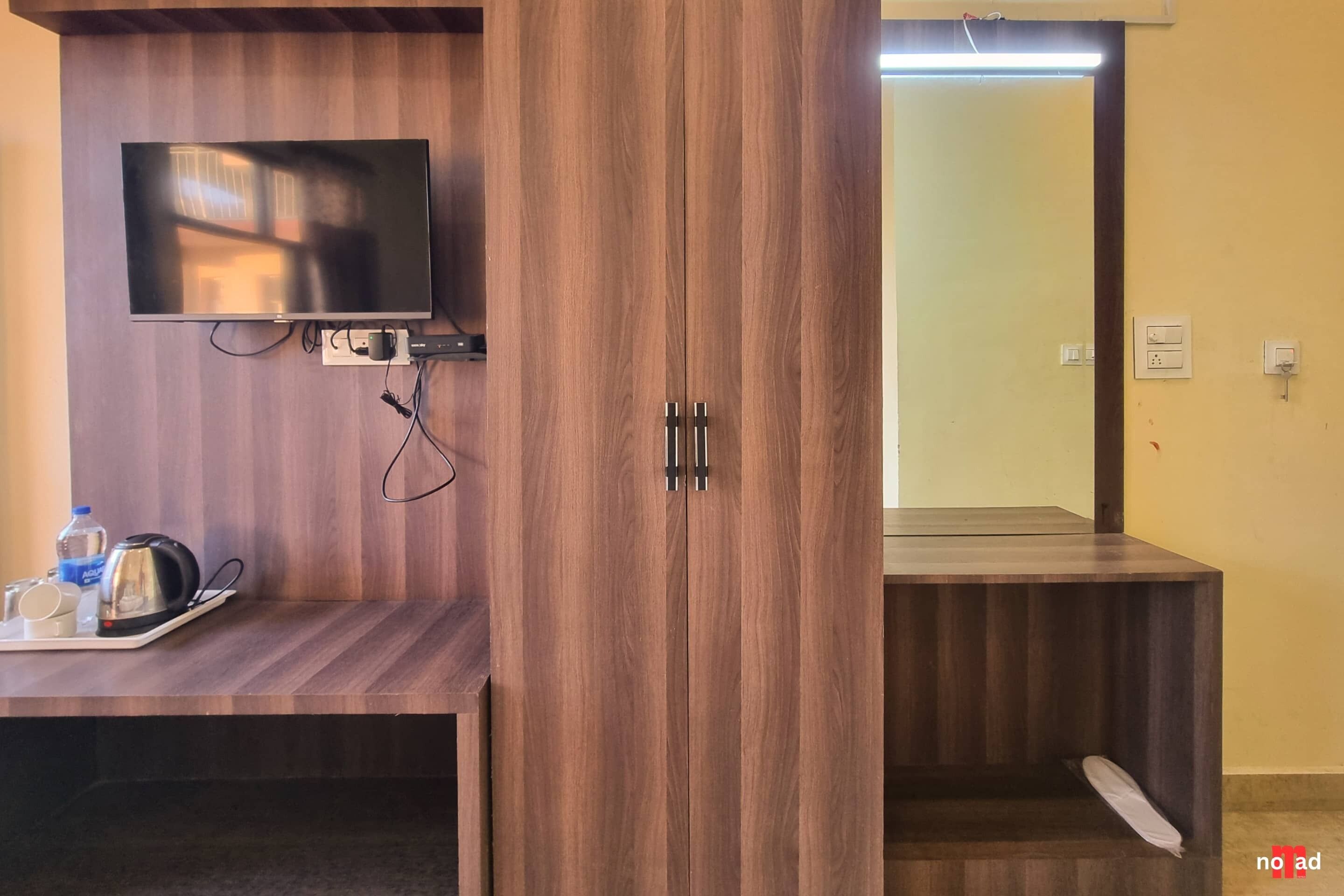 Quarto com guarda-roupa de madeira, televisão de tela plana na parede, mesa pequena com chaleira e chávenas, e espelho na porta do guarda-roupa.