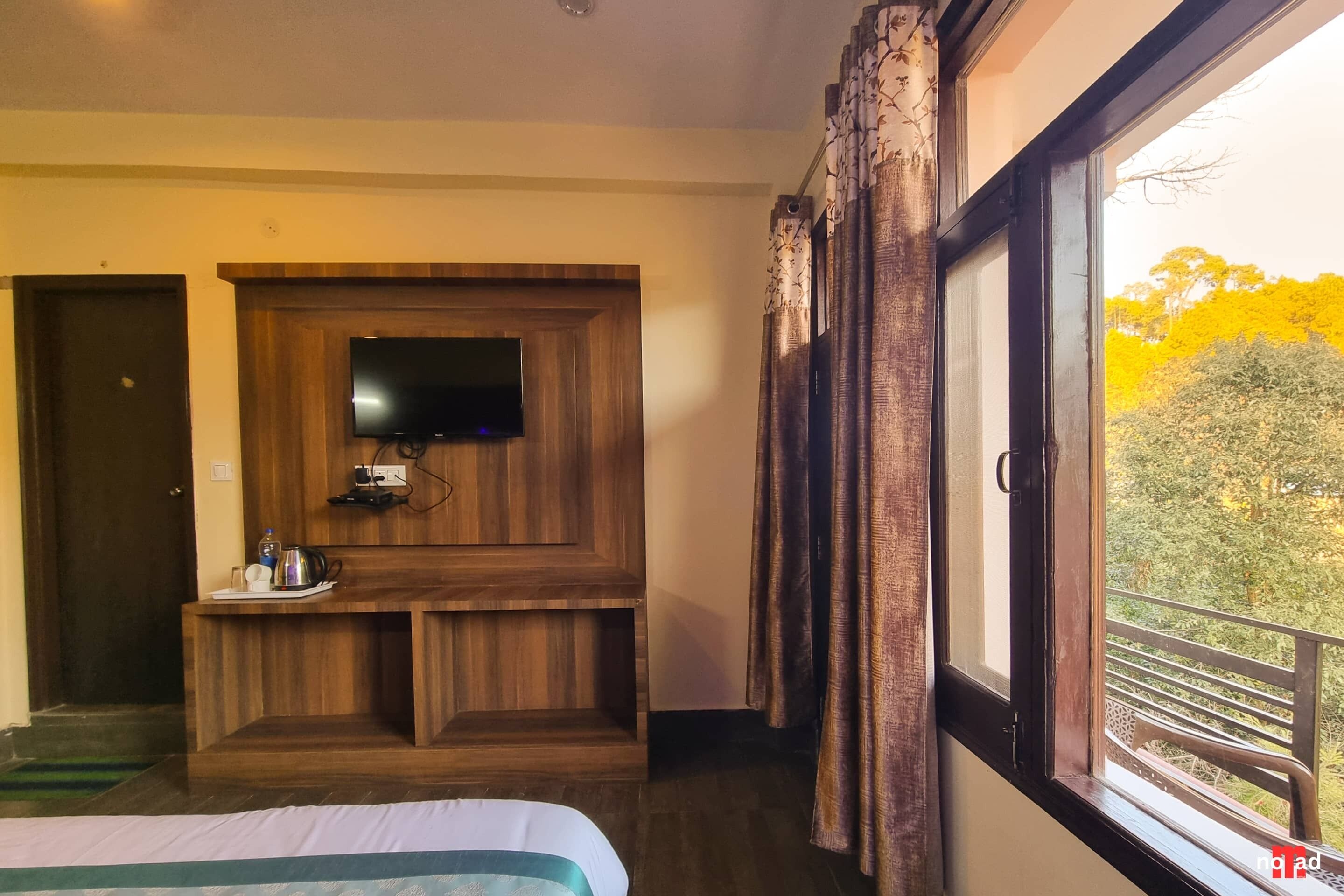 Habitación de hotel con un mueble de entretenimiento de madera, un televisor de pantalla plana, una mesita con una tetera y tazas, y un ventanal con vistas a árboles.