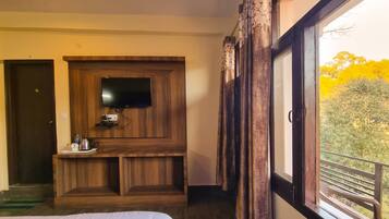 Habitación de hotel con un mueble de entretenimiento de madera, un televisor de pantalla plana, una mesita con una tetera y tazas, y un ventanal con vistas a árboles.