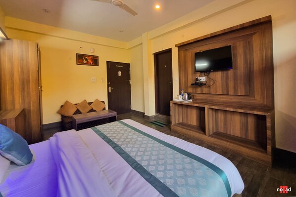 A hotel room with a bed, a wooden wardrobe, a television mounted on a wooden panel, and a bench with cushions.