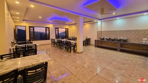 A spacious dining area with marble tables and chairs, a kitchen counter with appliances, and large windows offering a view of the landscape. - Nature Nest By Nomad Hotels (Dalhousie)