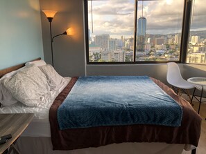 Ein Schlafzimmer mit Bett, Nachttisch, Lampe, Stuhl und Blick auf eine Stadtlandschaft durch große Fenster.