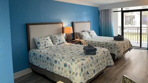 Ein Hotelzimmer mit zwei Betten, jedes mit einer gemusterten Tagesdecke, einem Nachttisch mit einer Lampe und Blick ins Freie durch ein großes Fenster.