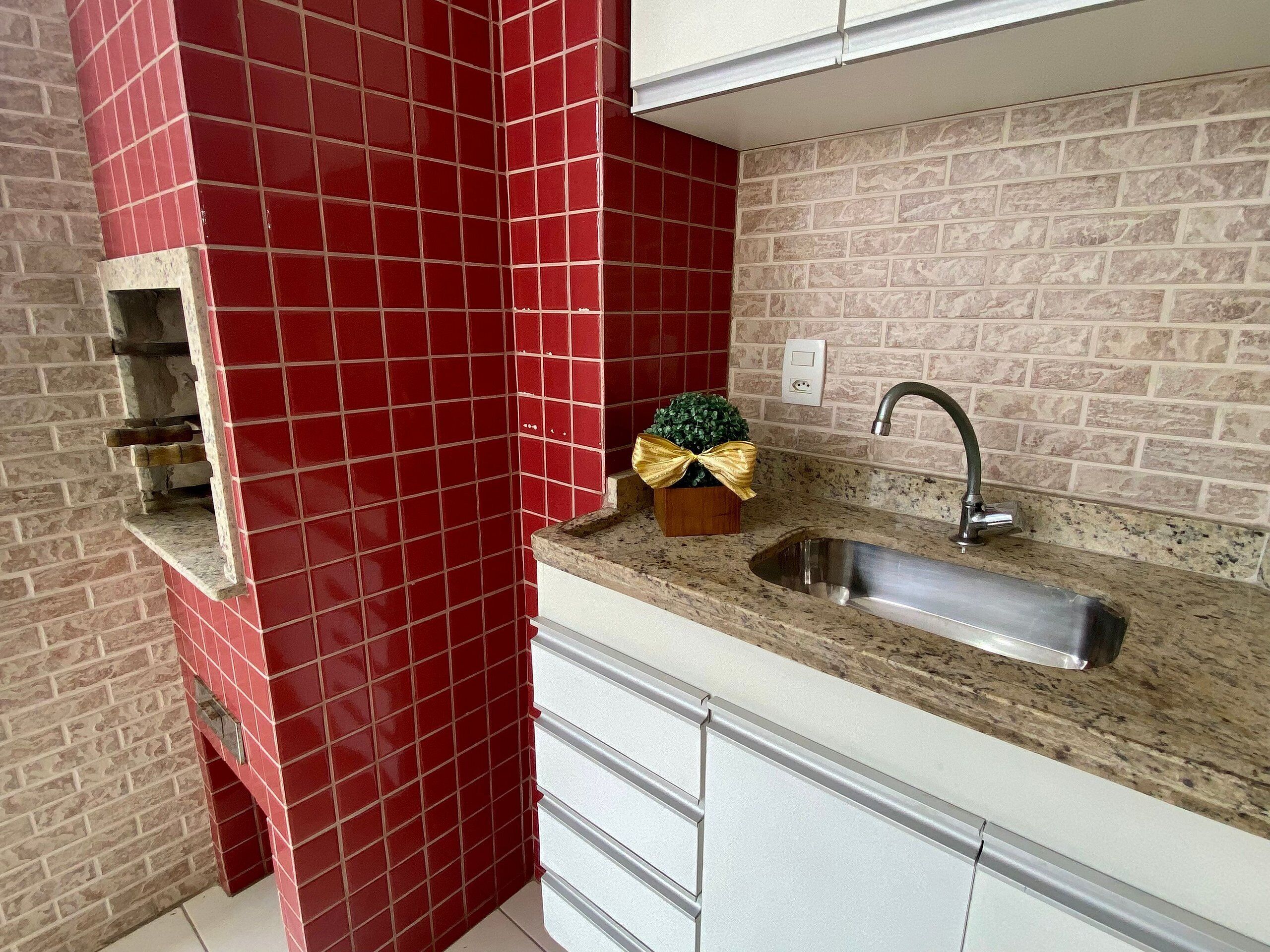 Une cuisine avec des murs recouverts de carreaux rouges, un plan de travail en granit, un évier en acier inoxydable et une petite boîte en bois ornée d’un ruban.