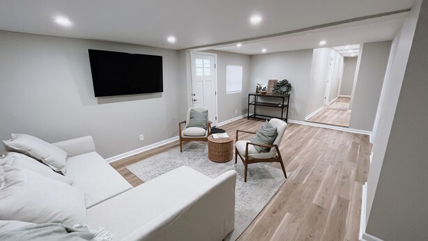 A spacious living room with a large flat-screen TV mounted on the wall, a white sofa, two armchairs, and a small round table.