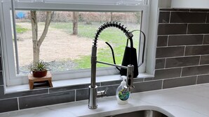 A kitchen with a stainless steel sink, a window with blinds, and a view of trees outside.