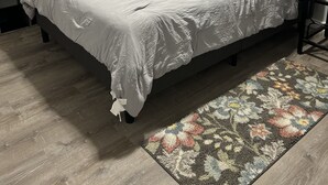 Un dormitorio con cama, mesita de noche, ventana con cortinas y alfombra estampada.