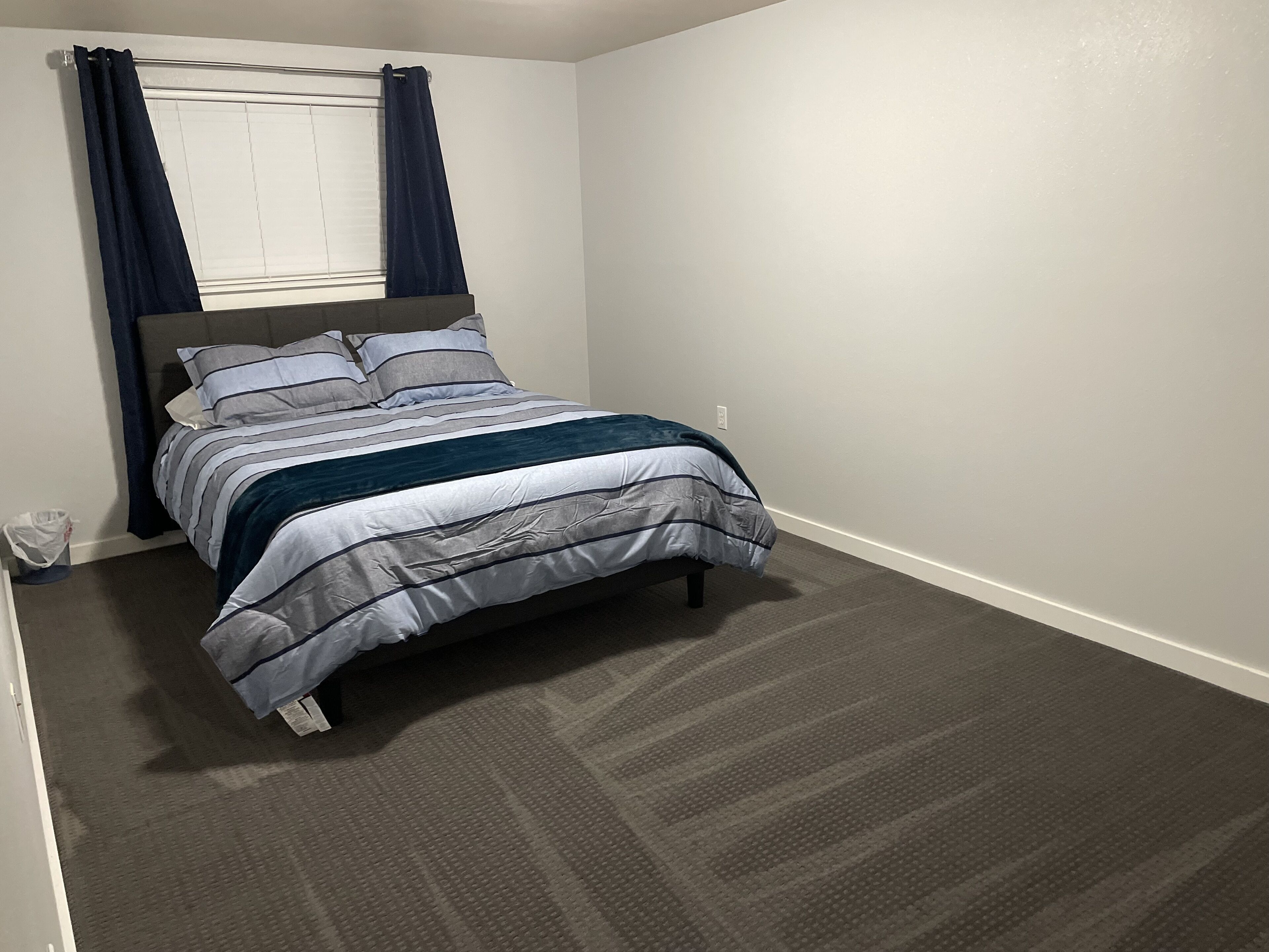A bedroom with a bed, dark curtains, a headboard, and a carpeted floor.