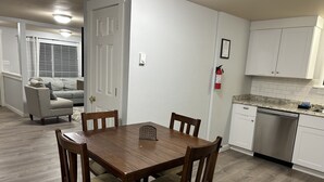 Un comedor con mesa y sillas de madera, una cocina con armarios blancos y lavavajillas, y una sala de estar con sofá y ventana con persianas.