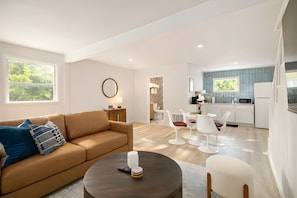 A modern living room with a sofa, coffee table, and white chairs. A kitchen is visible in the background with white cabinets and a window. - Melrose River Club (San Marcos)