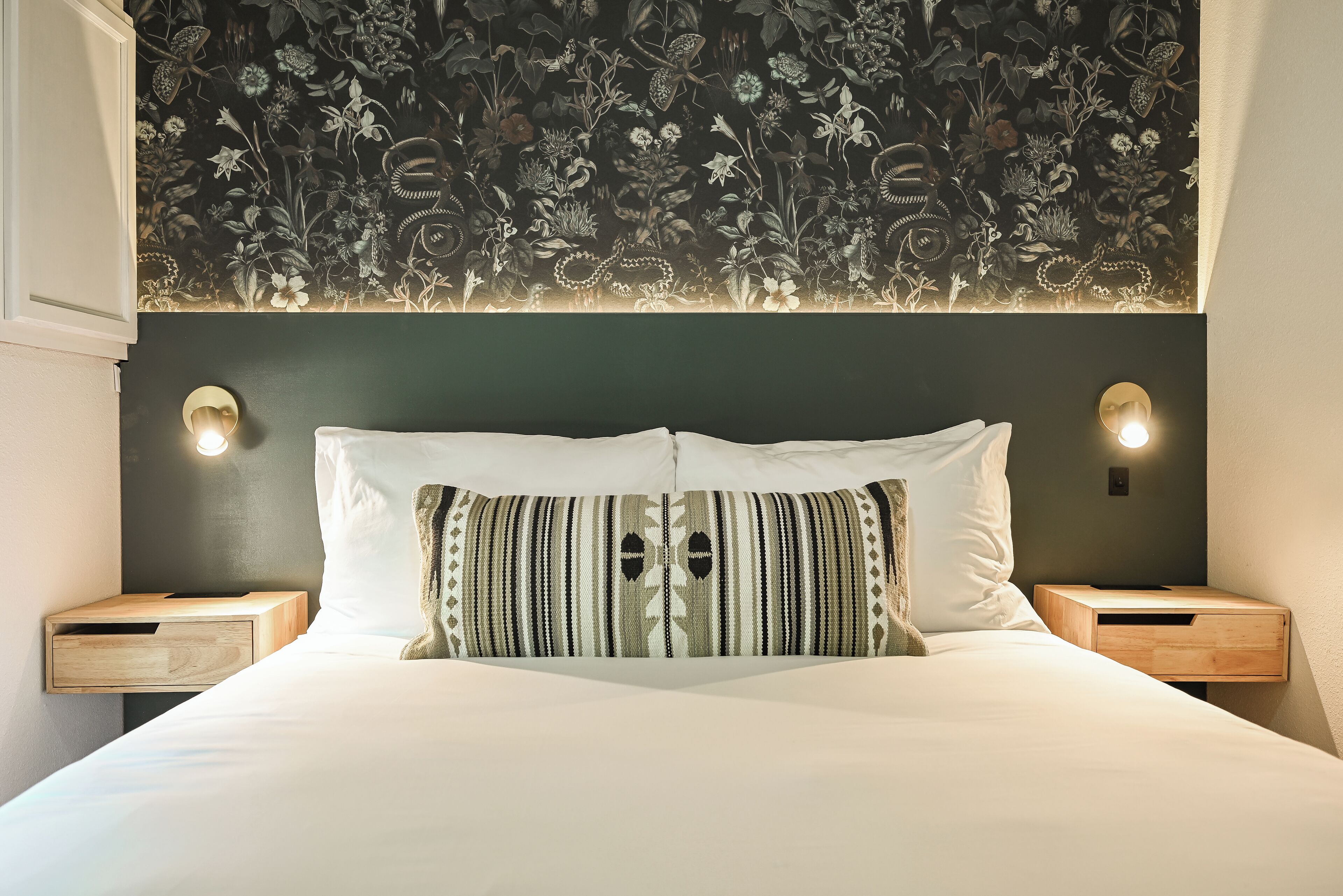 A neatly made bed with a patterned pillow, two bedside tables with lamps, and a floral wallpaper design on the wall above.