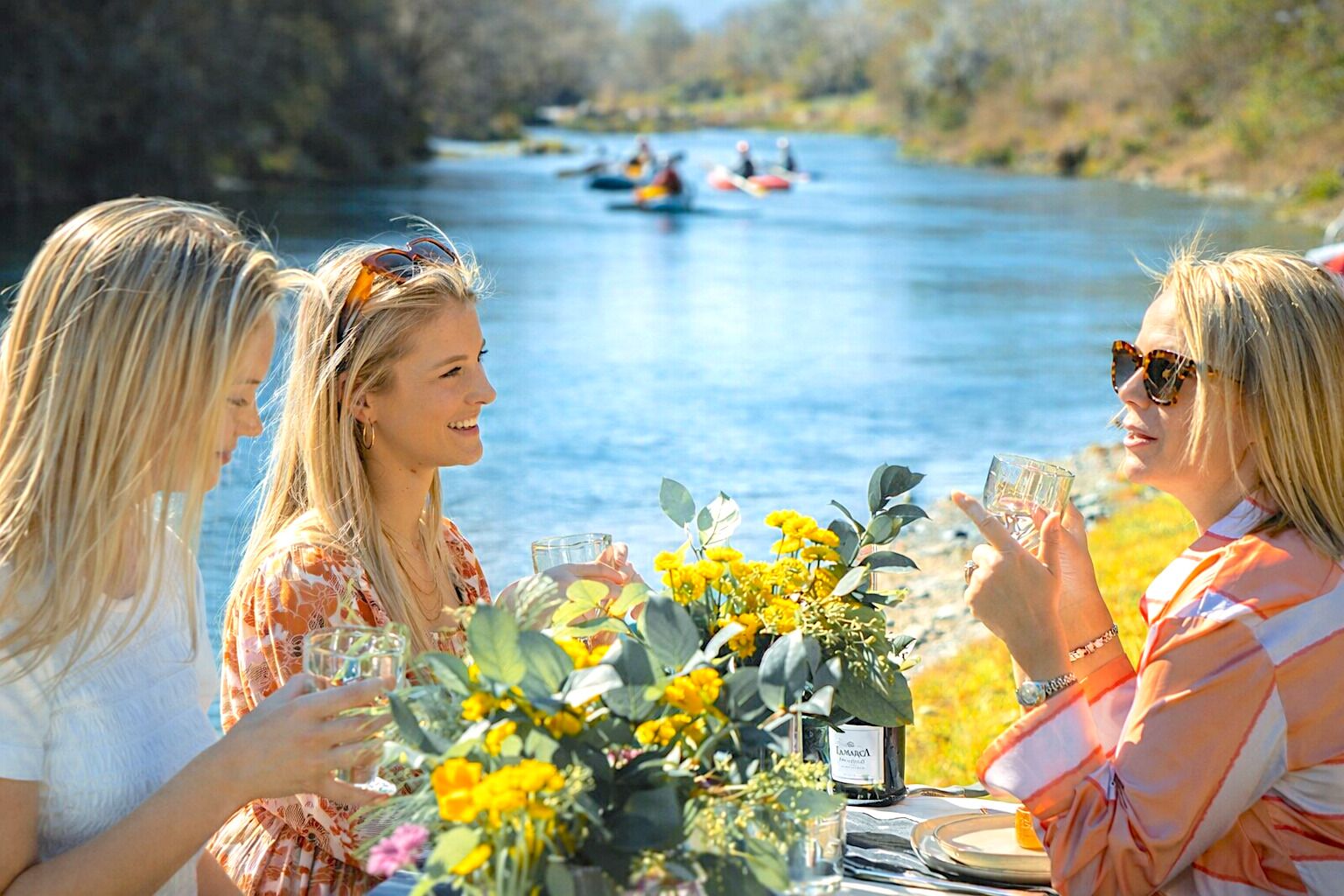 A group of people at a table with drinks and flowers, overlooking a river with kayaks in the background.