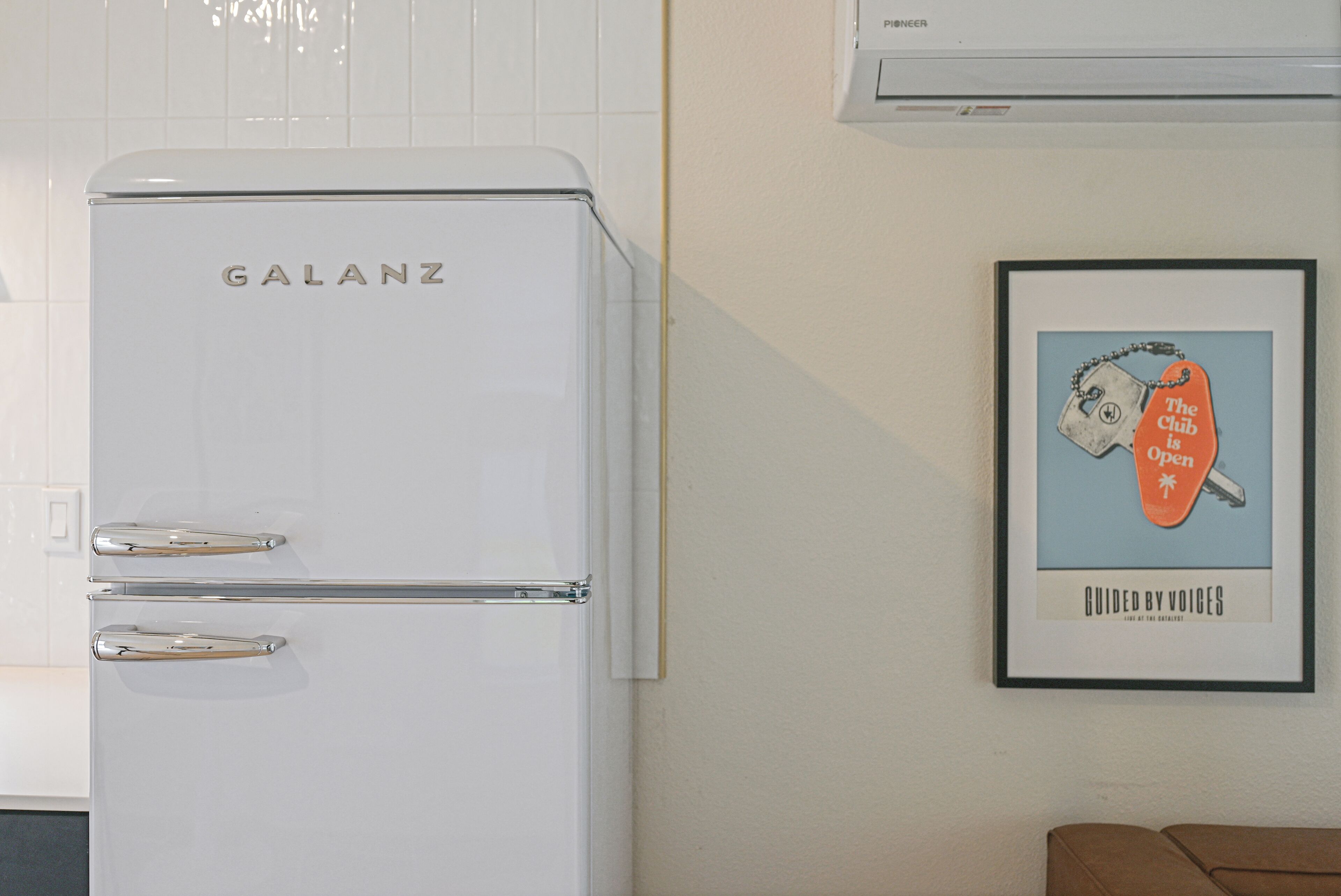 A white Galanz refrigerator in a kitchen with white tiled walls and a framed poster on the wall.