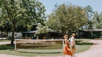 Señal con el texto 'MEET ME AT THE RIVER', rodeada de árboles y vehículos recreativos.