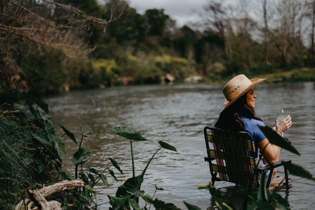 A person sitting on a chair by a river, holding a drink.