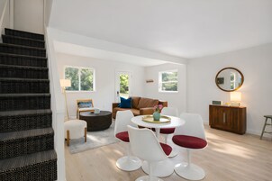 A modern living room with a staircase, a brown leather sofa, white chairs, a round table, and a wooden cabinet.