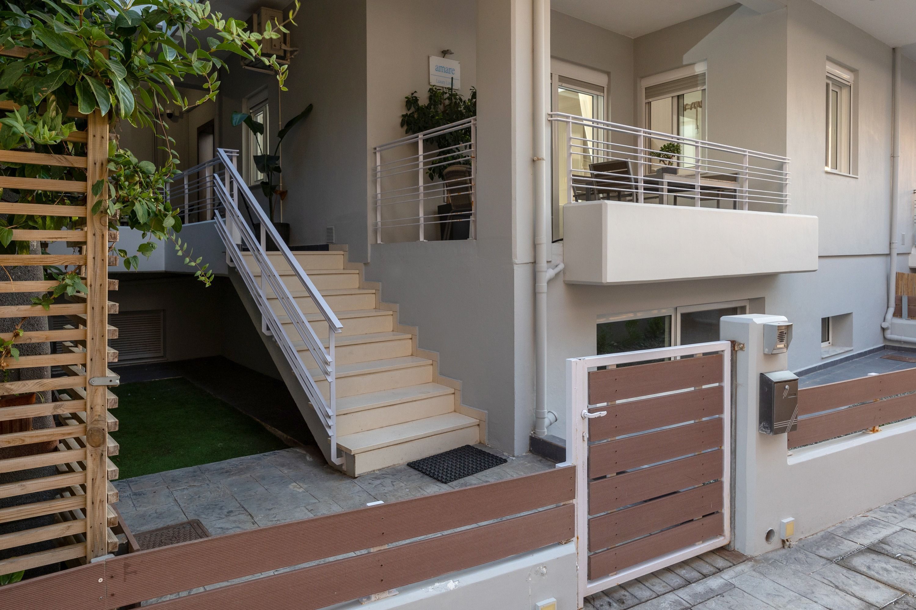 Un immeuble d’appartements moderne avec un escalier en bois menant à un balcon, une entrée sécurisée et un petit jardin.