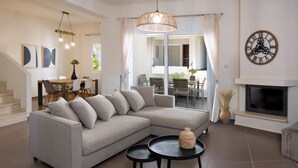 A modern living room with a sectional sofa, a dining area with a table and chairs, a fireplace, and a large wall clock. - Amare Luxury Living Volos (Volos)
