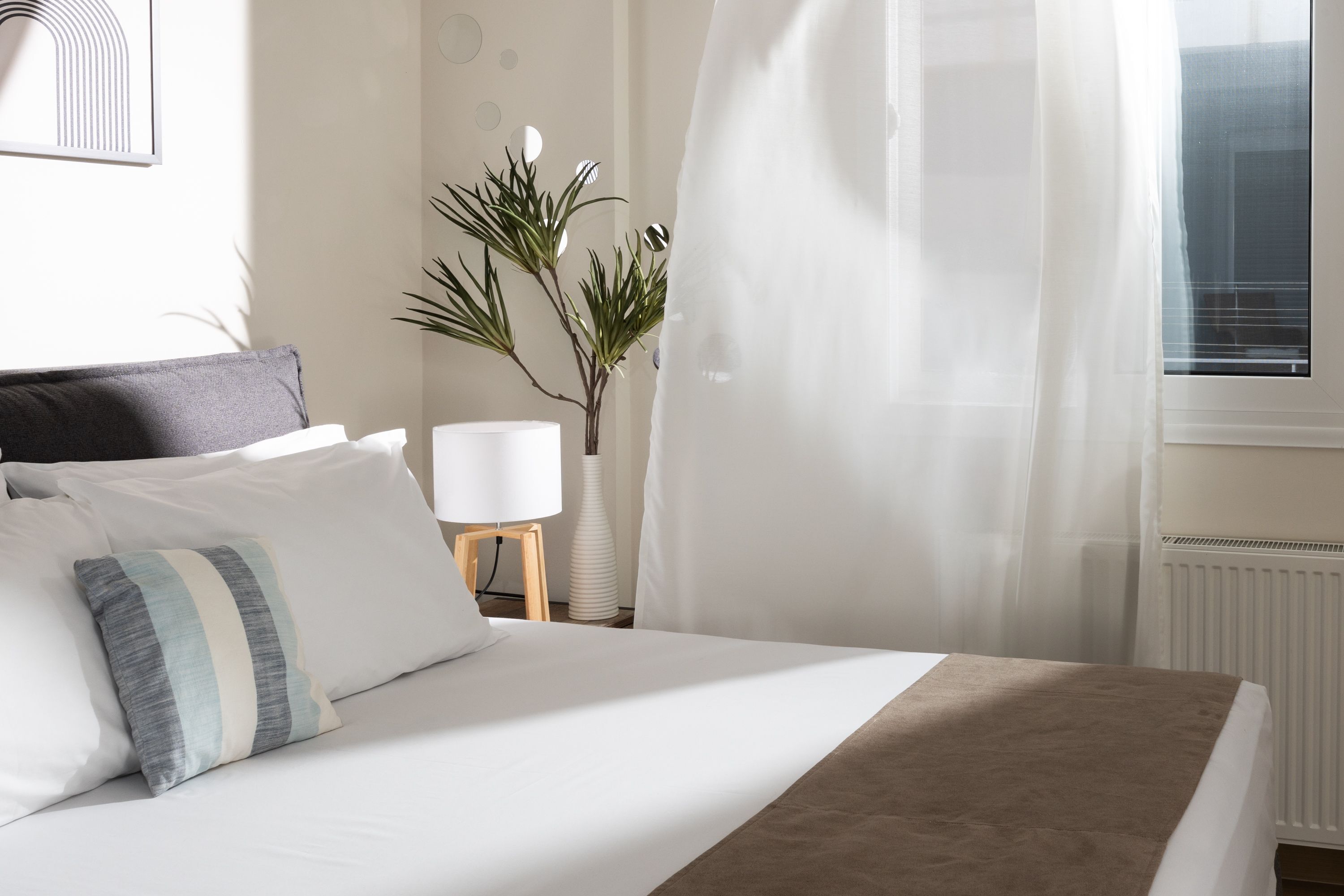 A neatly made bed with white and striped pillows, a bedside lamp, and a plant.