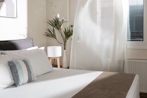 A neatly made bed with white and striped pillows, a bedside lamp, and a plant. - Amare Luxury Living Volos (Volos)