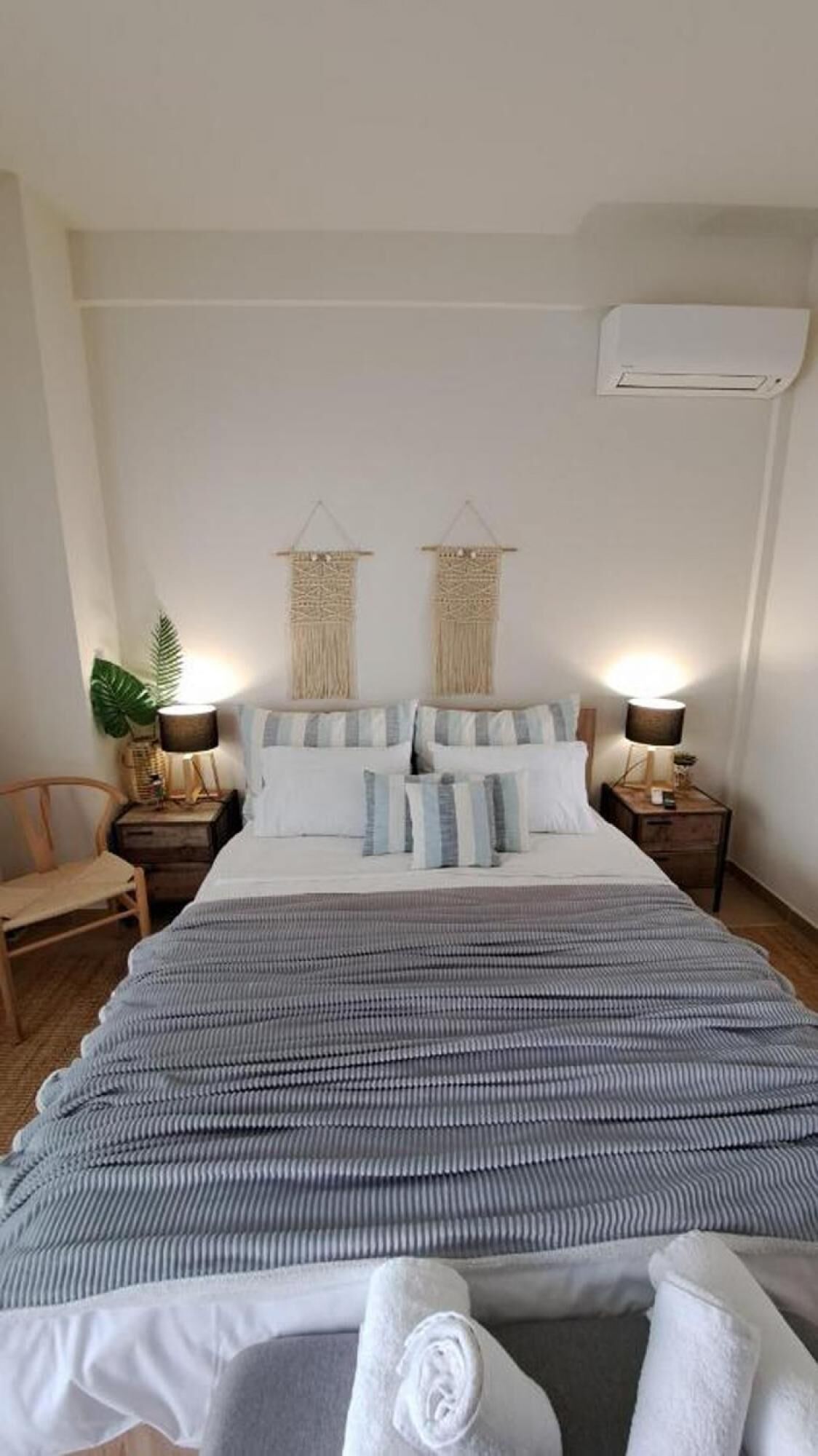A neatly made bed with striped bedding, two wall-mounted macramé hangings, and a small bedside table with a lamp on each side.