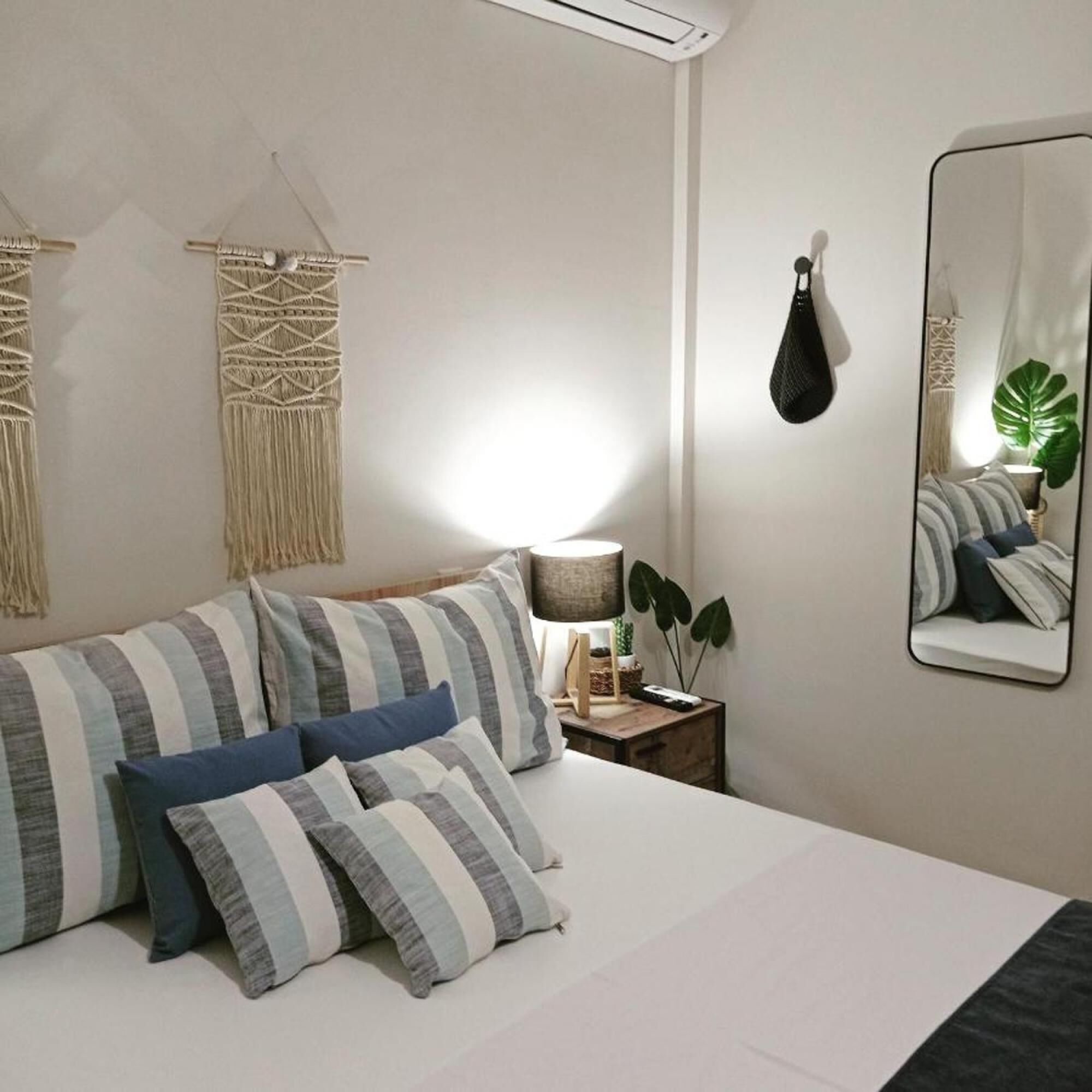 A neatly arranged bedroom with a bed, pillows, a bedside table with a lamp, a mirror, and macramé wall hangings.
