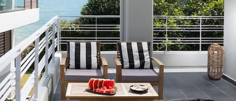 A balcony with a wooden sofa and cushions, a table with watermelon slices, and a view of the sea.