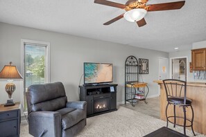 Una sala de estar con sofá de cuero, televisor de pantalla plana, chimenea, ventilador de techo y una zona de cocina con taburete de bar.