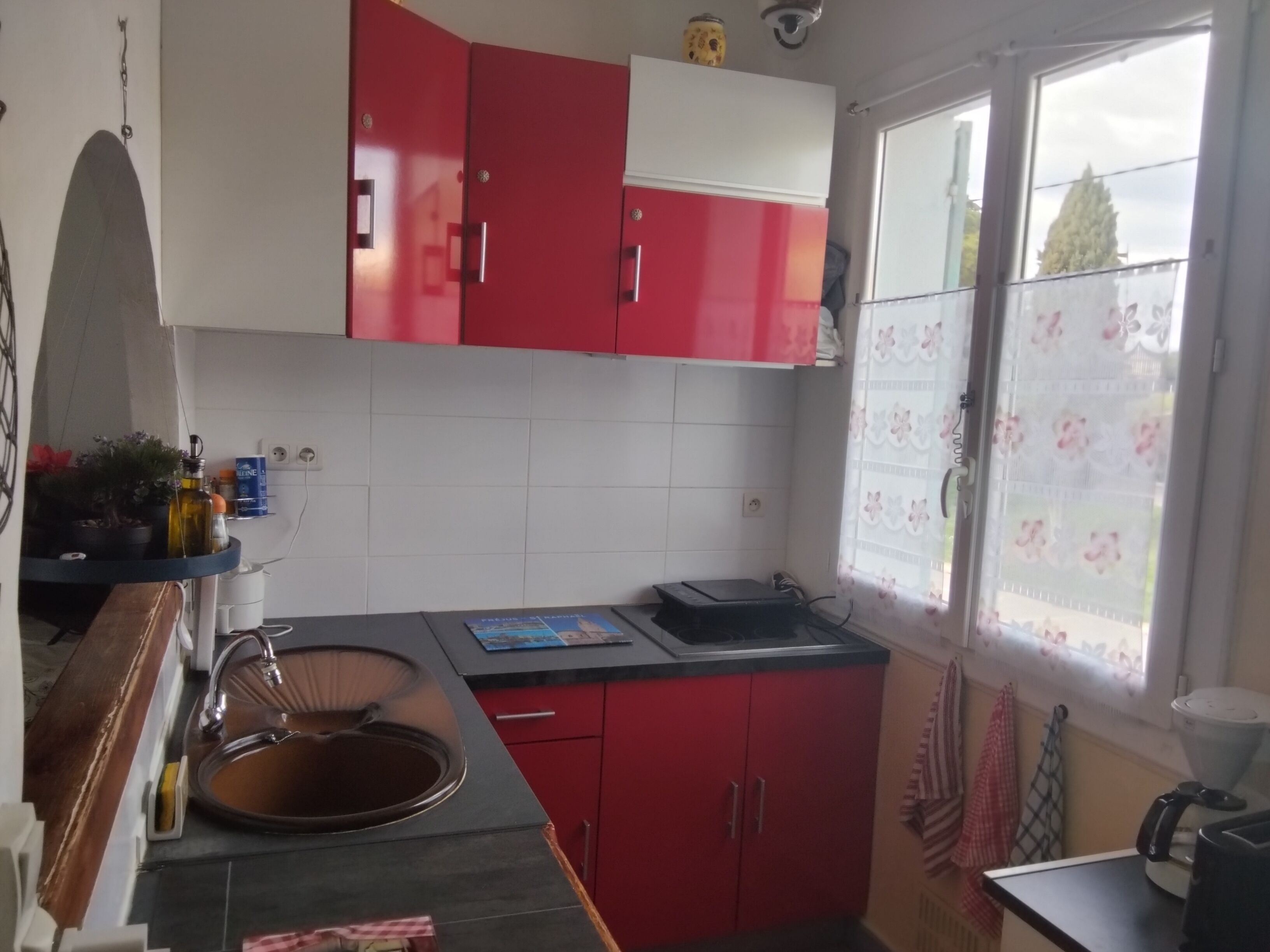 Une cuisine avec des meubles rouges et blancs, un évier, une fenêtre avec des stores à motifs floraux et un plan de travail sur lequel se trouvent une bouilloire et un petit appareil électroménager.