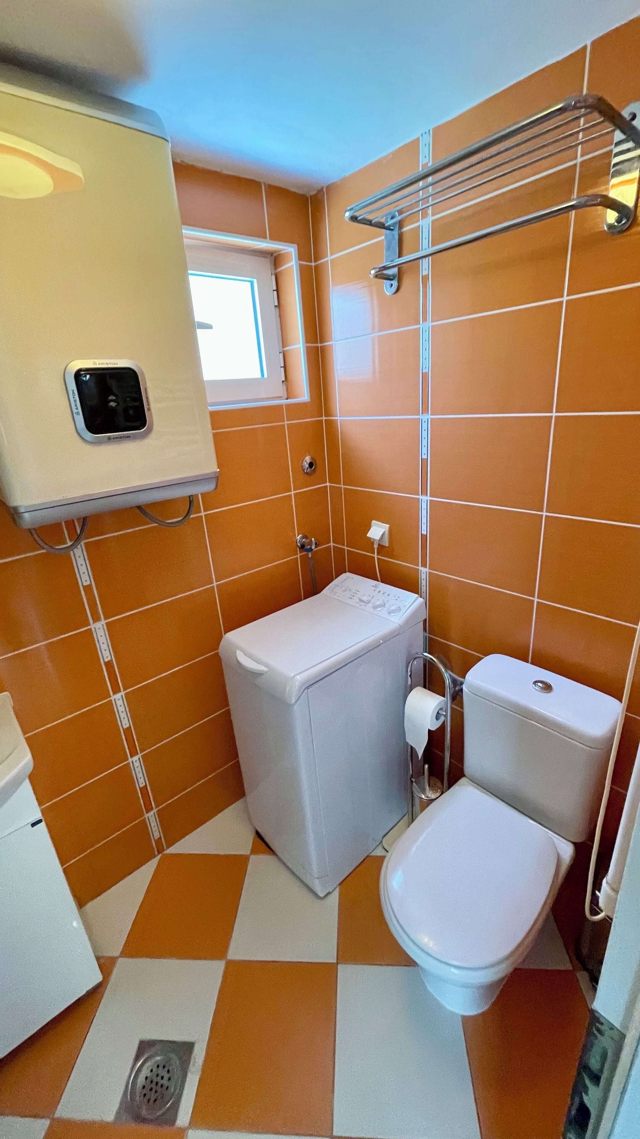 A compact bathroom with orange tiles, a white toilet, a washing machine, and a small window.