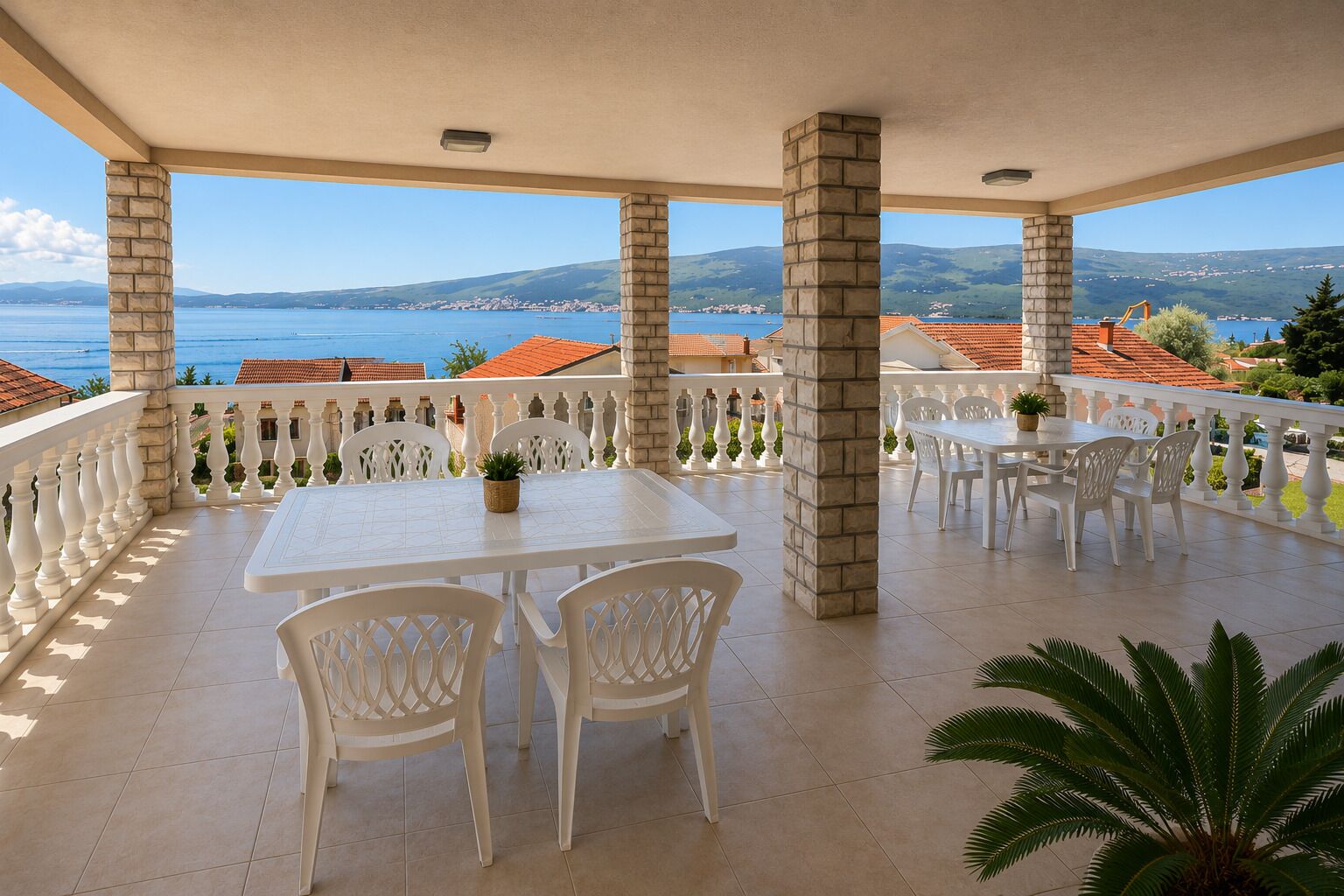 A covered outdoor area with a table and chairs, a white railing, and a view of the sea and buildings.