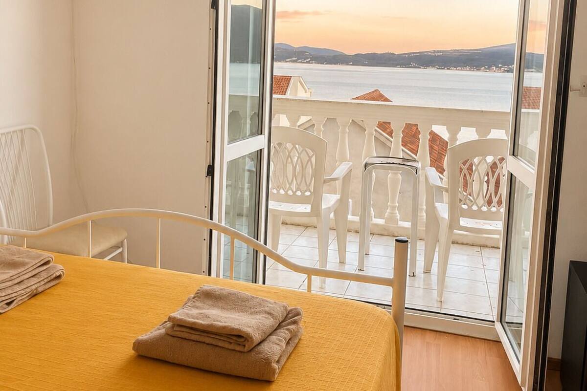 Una habitación con cama, una mesa con toallas, televisión y un balcón con sillas blancas y vista al mar.